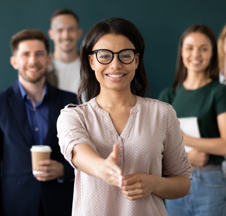 Mixed race woman sales manager stretch out hand introduces herself greeting shake hands client smiling look at camera pose indoors with diverse teammates. HR, job interview, business etiquette concept
